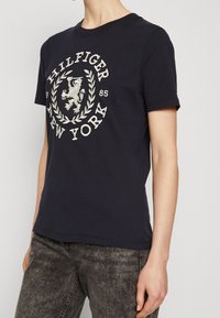 Mørkeblå bomulds t-shirt med et cirkulært broderet logo med en løve og laurbærdesign, rund hals og korte ærmer.