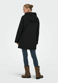 Duga crna parka s kapuljačom, s ravnim krojem i glatkom teksturom. Nosena preko plavih traperica i smešnih čizama do gležnja s kopčama.