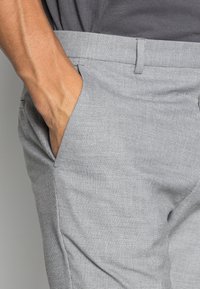 La mano di un uomo è appoggiata all'interno della tasca anteriore di pantaloni eleganti grigio chiaro con texture, indossando una camicia grigio scuro.