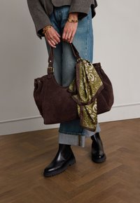 Bruine suède handtas met een groene geprinte sjaal vastgebonden aan het handvat, gecombineerd met blauwe spijkerbroeken en zwarte enkellaarsjes, neergezet op een houten vloer.