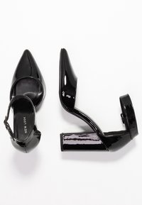 Chaussures à talons hauts en cuir verni noir avec un bout pointu, une bride de cheville et un talon épais. Texture lisse avec une finition brillante.