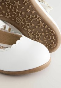 Zapatos blancos para niño pequeño con bordes ondulados y suelas de goma marrón grabadas con patrones de estrellas y el número seis.