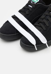 Mustat suleedist sneakerid lame mustade paeltega ja kahe valge paelaga, mis ristuvad ülaosas. Omab kummist tallat, millel on tekstuurne haard.
