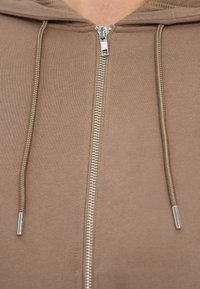 Κοντινό πλάνο ενός μπεζ φούτερ με φερμουάρ, με ασημί φερμουάρ και κορδόνια με μεταλλικές άκρες.