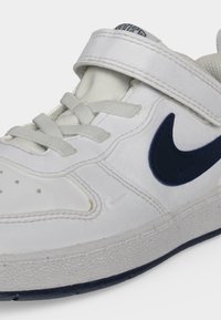 Λευκό αθλητικό παπούτσι Nike με ναυτικό σήμα, λουράκι με βέλκρο, λευκά κορδόνια και υφή στη λαστιχένια σόλα, σε κοντινή λήψη.