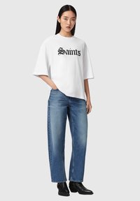Giovane donna che indossa una t-shirt bianca oversize con la scritta "Saints", jeans blu a gamba larga e stivaletti neri, in piedi con una mano in tasca.