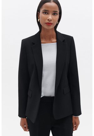 Blazer noir au design ajusté, avec des revers en pointe et deux poches avant. Porté par-dessus un haut gris clair, présentant une apparence nette et professionnelle.