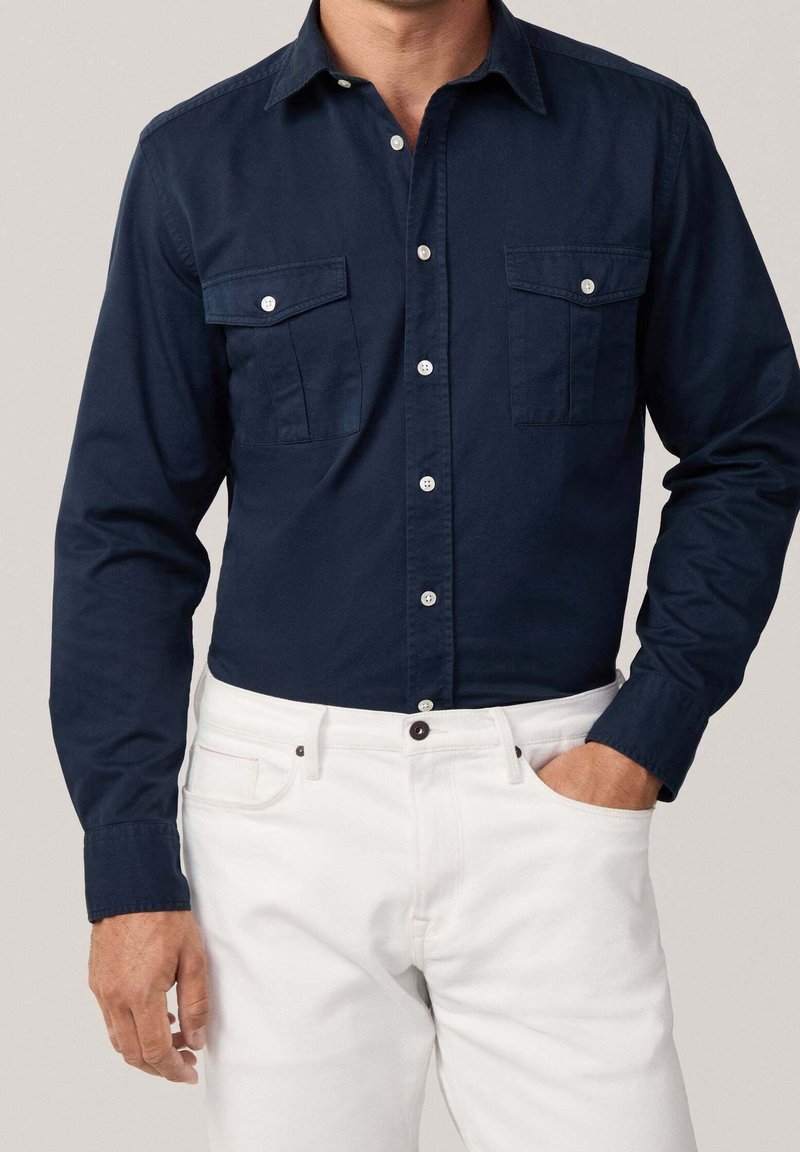 Marineblaues Langarmhemd mit Button-Down-Kragen, zwei Brusttaschen und weißen Knöpfen, kombiniert mit weißen Hosen. Glatte Stofftextur.