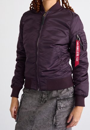 Personne portant une veste bomber violet foncé avec des poignets et un ourlet côtelés, une fermeture éclair à l'avant, et une étiquette rouge "Remove Before Flight" sur la poche de la manche gauche.