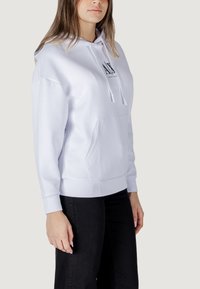 Sudadera blanca hecha de tela suave, con un bolsillo frontal, cordones y un logo impreso en el pecho. Acompañada de pantalones negros.
