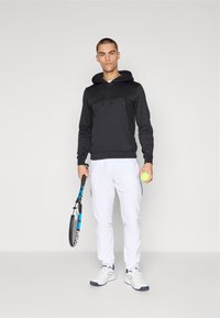 Schwarzer Kapuzenpullover mit einer Fronttasche, kombiniert mit weißen Sporthosen. Das Model hält einen blauen Tennisschläger und einen gelben Tennisball.