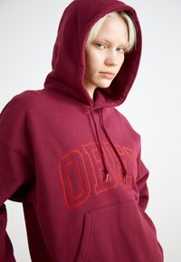 Μπορντό φούτερ με κουκούλα, με το λογότυπο "OBEY" κεντημένο σε πορτοκαλί χρώμα; διαθέτει τσέπη μπροστά και κουκούλα με κορδόνια. Μαλακό, υφασμάτινο υλικό.