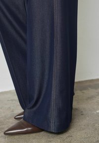 Széles szárú tengerészkék nadrág, sima anyagból, kontrasztos, világos varrással a varratok mentén, hegyes barna bőrcipővel.