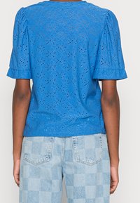 Blusa azul con ojales y mangas abullonadas, de escote redondo. Presenta una textura estampada con cortes. Combinada con unos vaqueros de cuadros en azul claro.
