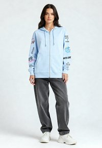 Felpa con cappuccio azzurro chiaro con zip e tasche frontali, con grafiche colorate e testo sulle maniche. Indossata con jeans grigi larghi e sneakers bianche.
