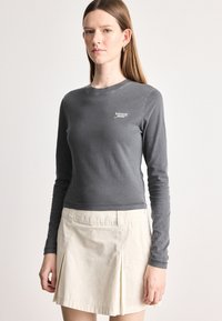 Γκρίζο πουκάμισο μακρυμάνικο με στρογγυλή λαιμόκοψη και λευκό λογότυπο "tommy jeans". Συνδυάζεται με μια ανοιχτόχρωμη μπεζ φούστα με πτυχώσεις. Λείος υφασμάτινος υφής.