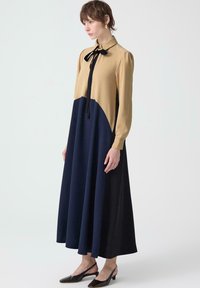 Femeie îmbrăcată cu o rochie lungă în două tonuri, cu un top cămilă, fustă bleu închis, papion negru la guler și pantofi slingback cu toc mic și negru.