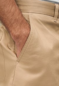 Main d'homme reposant dans la poche avant d'un pantalon beige avec une boucle de ceinture visible et une texture de tissu.