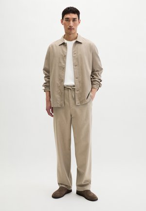 Mann trägt beige locker sitzende Jacke und Hose über weißem Hemd mit braunen Slippern, steht vor hellem einfarbigem Hintergrund.