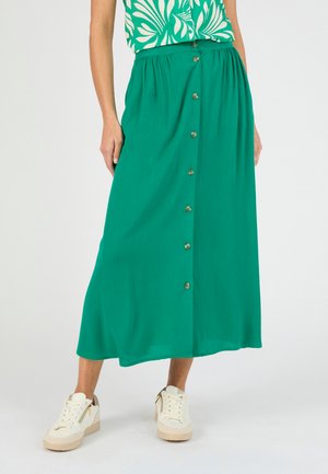 Vrouw met een groene midirok met knopen aan de voorkant, witte sneakers en een groen patroon topje, ingestopt, staand tegen een neutrale achtergrond.