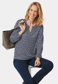 Marineblouse met wit geometrisch patroon, v-hals en lange gathered mouwen, gecombineerd met donkere jeans en een bruine schoudertas.