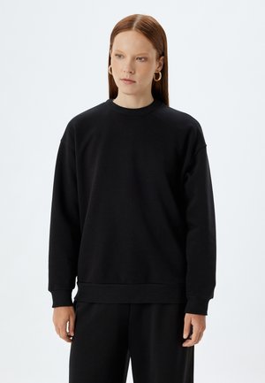 Zwarte sweatshirt van zacht materiaal; voorzien van een ronde hals, verlaagde schouders en geribbelde manchetten. Gecombineerd met bijpassende zwarte broeken.