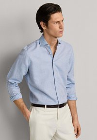 Uomo con camicia azzurra chiusa con bottoni e maniche arrotolate, pantaloni bianchi, in piedi con una mano nella tasca, che guarda verso destra.