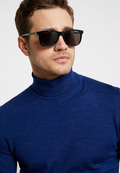 Schwarze Sonnenbrille mit dunklen Gläsern und rechteckigem Rahmen, getragen mit einem enganliegenden blauen Rollkragenpullover, der eine glatte, weiche Textur zeigt.
