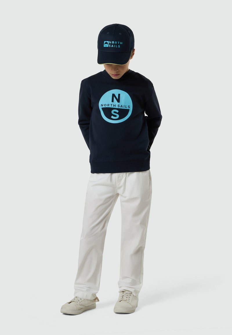 Bambino che indossa un cappellino North Sails blu navy e una felpa con logo, pantaloni bianchi e sneakers beige in piedi davanti a uno sfondo semplice.