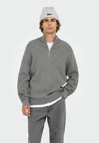 Grauer Zip-Hals-Pullover mit gerippter Textur, langen Ärmeln und einem lockeren Schnitt. Kombiniert mit hellgrauen Jeans und einer hellgrauen gerippten Mütze.