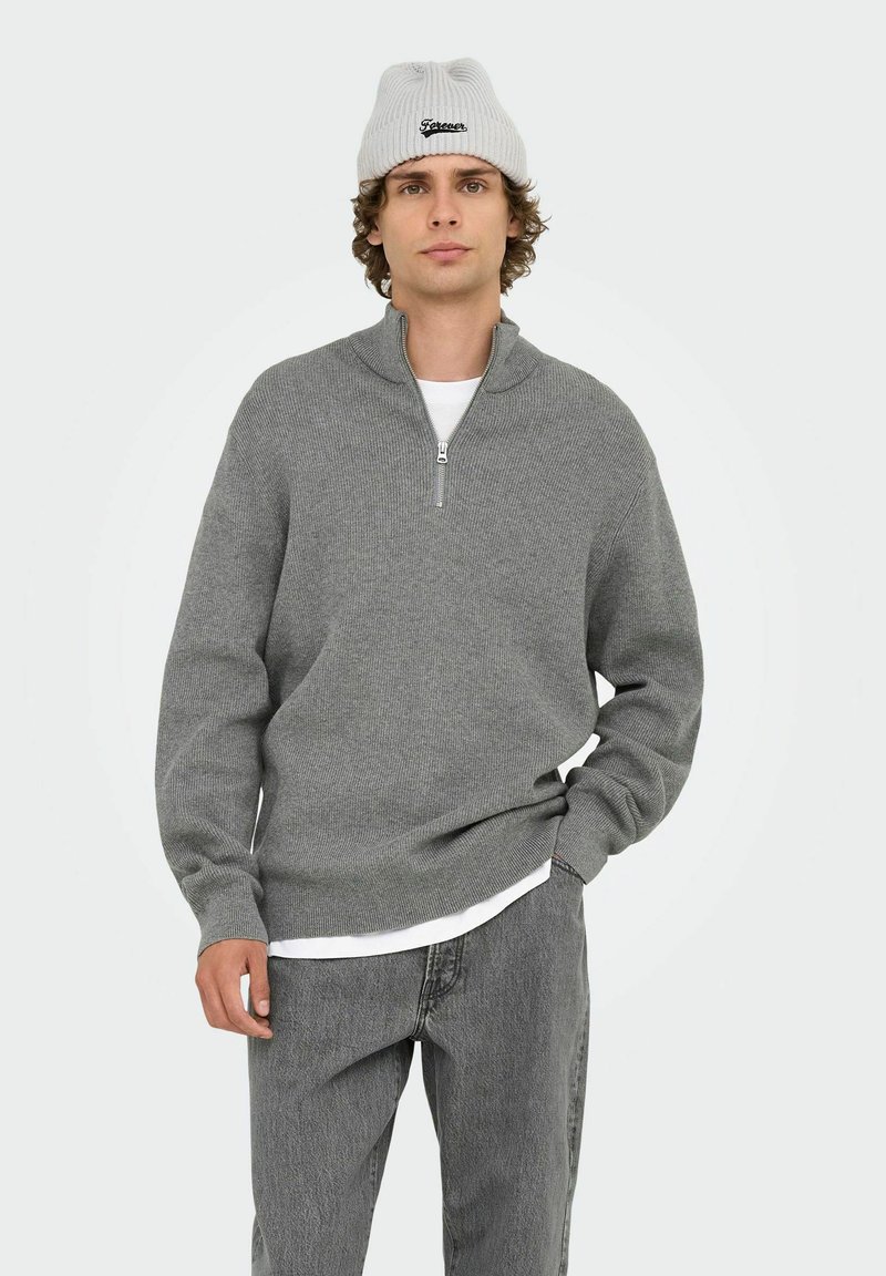 Grauer Zip-Hals-Pullover mit gerippter Textur, langen Ärmeln und einem lockeren Schnitt. Kombiniert mit hellgrauen Jeans und einer hellgrauen gerippten Mütze.