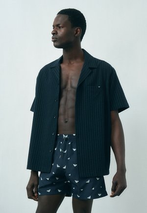 Homme à la peau foncée portant une chemise noire à manches courtes ouverte et un short de bain bleu marine avec des logos blancs, debout devant un fond clair uni.