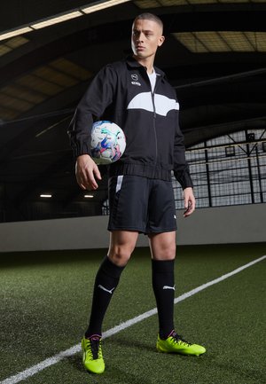 Chaqueta atlética negra con acentos blancos, pantalones cortos a juego, calcetines negros y zapatillas brillantes verdes. Pelota sostenida en la mano izquierda, de pie sobre césped.