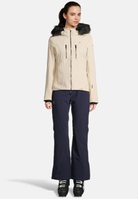 Damen Skijacke in Creme mit Kapuze mit Fellbesatz, mit Reißverschlusstaschen, kombiniert mit marineblauen Skihosen und schwarzen Skistiefeln. Glatte Textur und taillierte Passform.