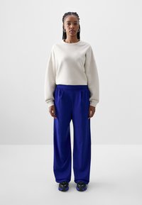 Krem majica s opuštenim ramenima, uparena s širokim kraljevsko plavim hlačama i crnim tenisicama s ljubičastim detaljima.