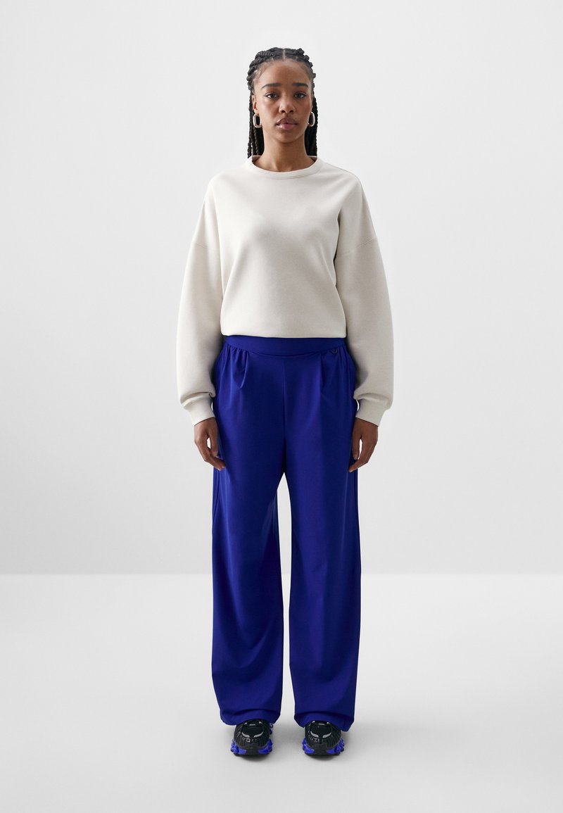 Krem majica s opuštenim ramenima, uparena s širokim kraljevsko plavim hlačama i crnim tenisicama s ljubičastim detaljima.