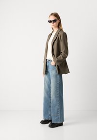 Υπερμεγέθης ελαιοπράσινος σακάκι, λευκό μπλουζάκι, ανοιχτό μπλε παντελόνι wide-leg και μαύρες chunky μπότες με τακούνι; μοντέλο φοράει μαύρα γυαλιά ηλίου.