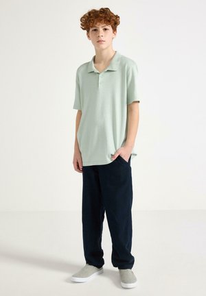 Punatukkainen nuori seisoo käsi taskussa, yllään vaaleanvihreä poolopaita, tummat housut ja vaaleat harmaat slip-on-kengät.