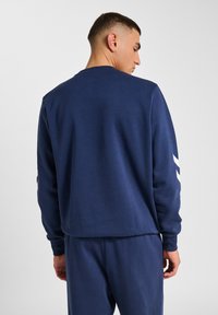 Man med kort hår iförd en marinblå långärmad sweatshirt och matchande byxor, vänd bort med ryggen mot kameran och vit zigzag på ärmen.