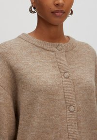 Beige gebreide cardigan met een ronde halslijn, ribbelige knoopsluiting en textuurstof. Voorzien van twee grote knopen en een zachte afwerking.