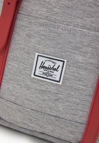 Grå tygväska med röda remmar, med en vit etikett som har texten "Herschel Supply Co." Strukturad yta med synliga sömmar.