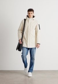 Beige vandtæt parka med hætte, front lynlås, sort patch på ærmet, parret med blå jeans og hvide sneakers, båret med en sort taske.