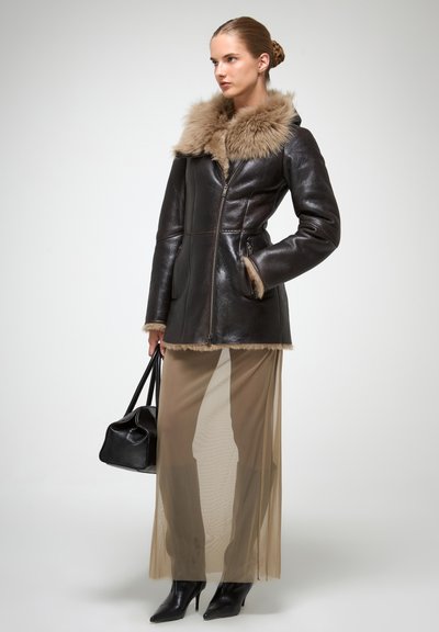 Brūns ādas jaka ar kažoka apkakli, priekšējais rāvējslēdzis un sānu kabatas, apvienota ar caurspīdīgu bēšu svārku un melnu šauru purngalu apavus.