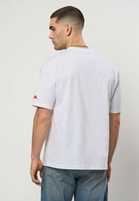 Άντρας με κοντά μαλλιά που φοράει απλό λευκό T-shirt με μικρό κόκκινο λογότυπο στο δεξί μανίκι και μπλε τζιν, γυρισμένος πλάτη στην κάμερα.