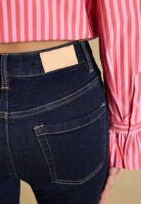 Personne portant un jean bleu foncé avec un patch en cuir beige et un t-shirt à manches longues rayé rose et rouge.