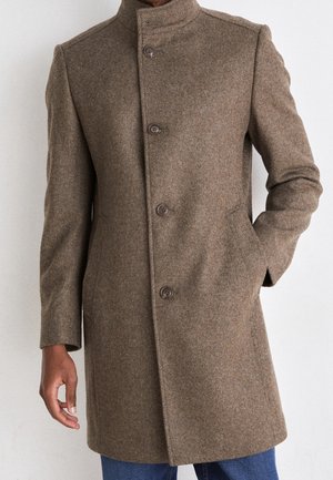 Homme portant un manteau en laine marron longueur genoux avec quatre boutons, debout la main dans la poche devant un fond uni.