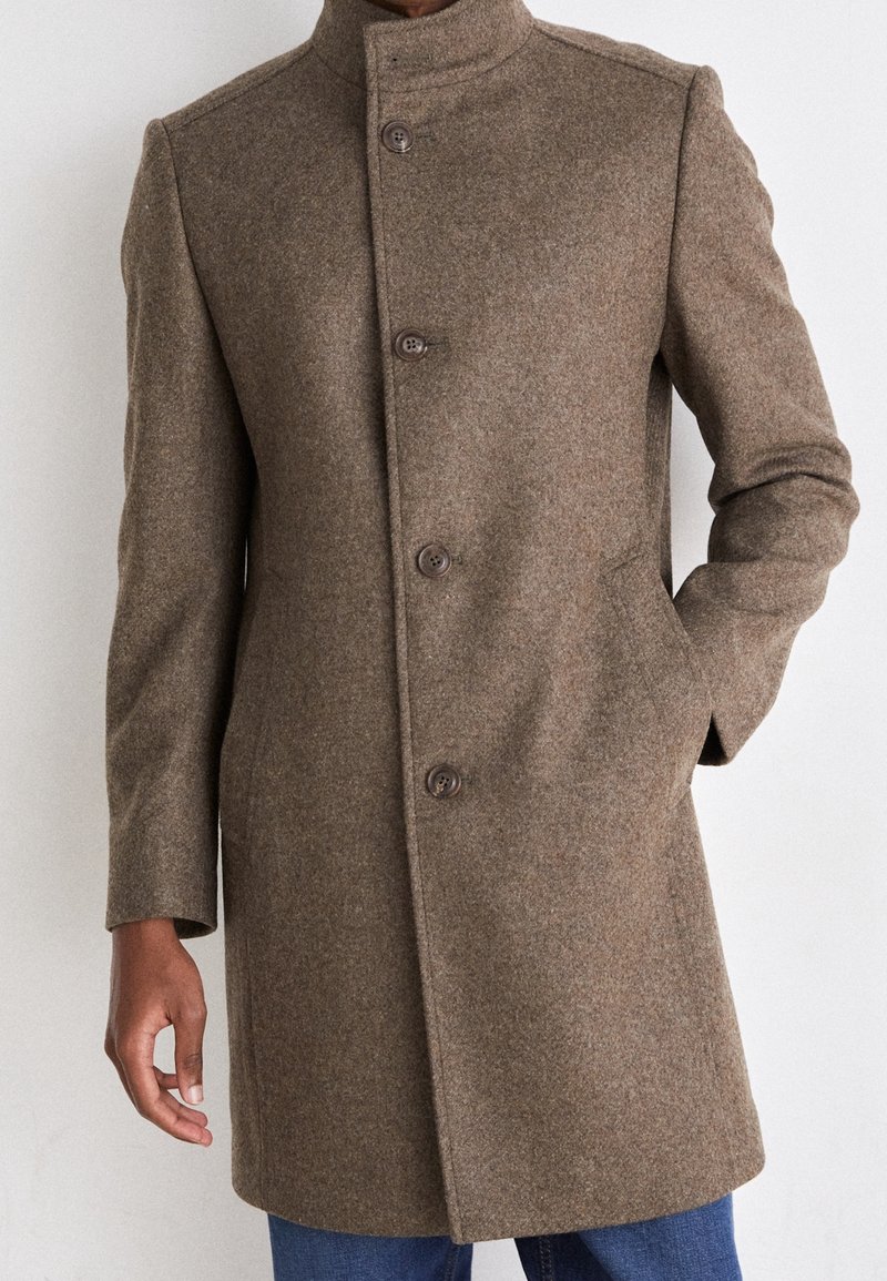 Homme portant un manteau en laine marron longueur genoux avec quatre boutons, debout la main dans la poche devant un fond uni.