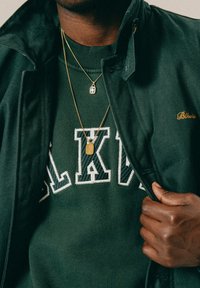 Άτομο που φοράει σκούρο πράσινο σακάκι και πουλόβερ με μερικά λευκά γράμματα "LAKA," κρατώντας ανοιχτό το σακάκι για να δείξει δύο χρυσές κολιέ.