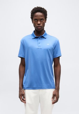 Fiatal férfi áll, kék rövid ujjú galléros ingben és fehér nadrágban, egyszerű világos háttér előtt.