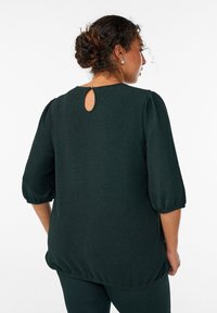Dunkelgrüne Bluse aus strukturiertem Stoff, runder Ausschnitt, Dreiviertelärmel und ein kleines Schlüsselloch-Detail an der Rückseite.